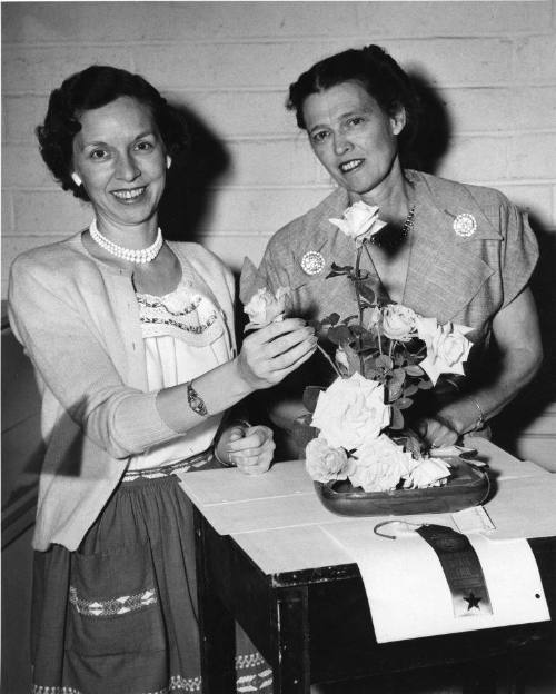 Two women stand behind a table holding a flower arrangement in the flower show