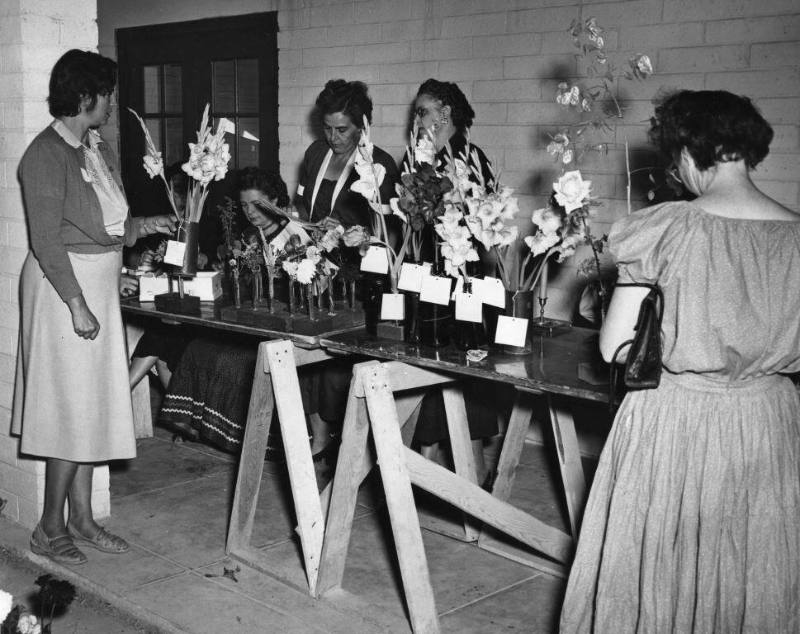 Flower Show competitors stand around a table full of flowers in vases