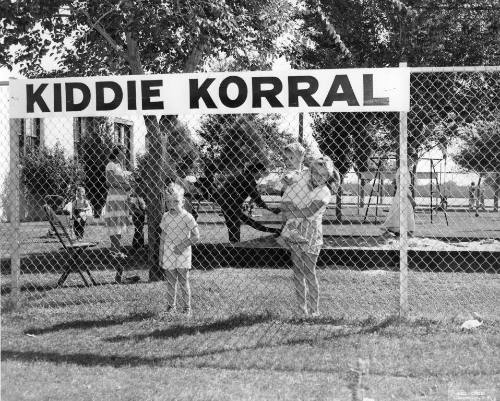 Children stand behind a chain link fence with the words "Kiddie Korral" on it
