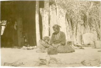 Native American woman and child sit outside a log building