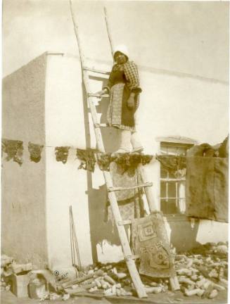 Native American woman stands on ladder outside of an adobe house