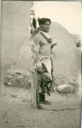 Native American man standing in front of an horno in dance attire
