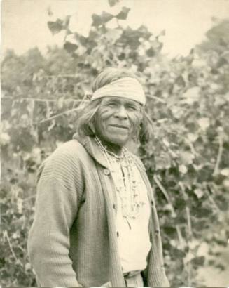 Older Native American man stands in front of vines, looking at the camera