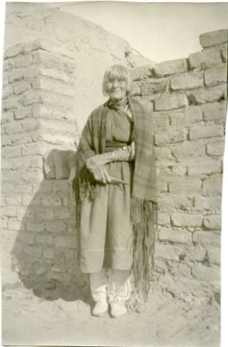 Older Native American woman standing beside an adobe wall, wearing a shawl