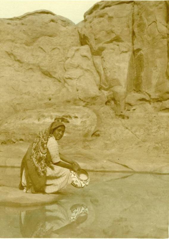An Acoma woman kneels next to a stream, holding a pot