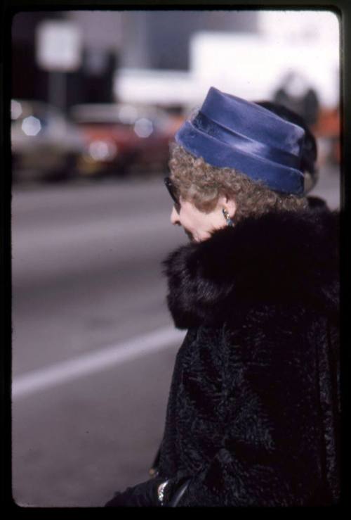 A woman in a fur coat and a blue velvet hat waits to cross a street