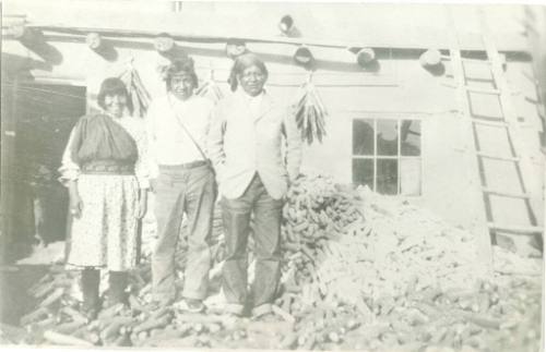 A woman and two men stand outside of a house with a large pile of corn