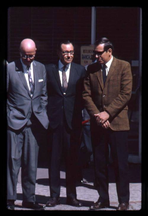 Three men in business suits stands on a sidewalk